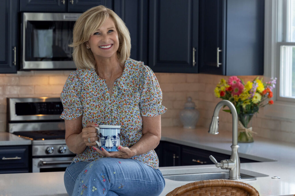 blonde woman smiling in a floral shirt while holding a coffee mug in the kitchen