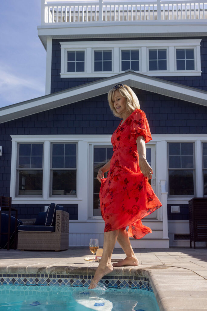 blonde woman in a red dress dipping her toes in the swimming pool with a blue house in the background