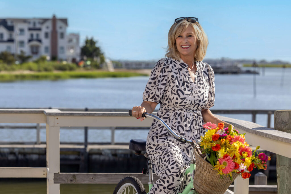 a blonde woman in a black and white print dress riding a green bike with flowers in the brown basket on a sunny day