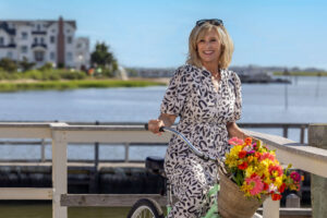 a blonde woman in a black and white print dress riding a green bike with flowers in the brown basket on a sunny day