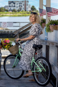 a blonde woman in a black and white dress riding a green bike on a bay dock with flowers in the basket
