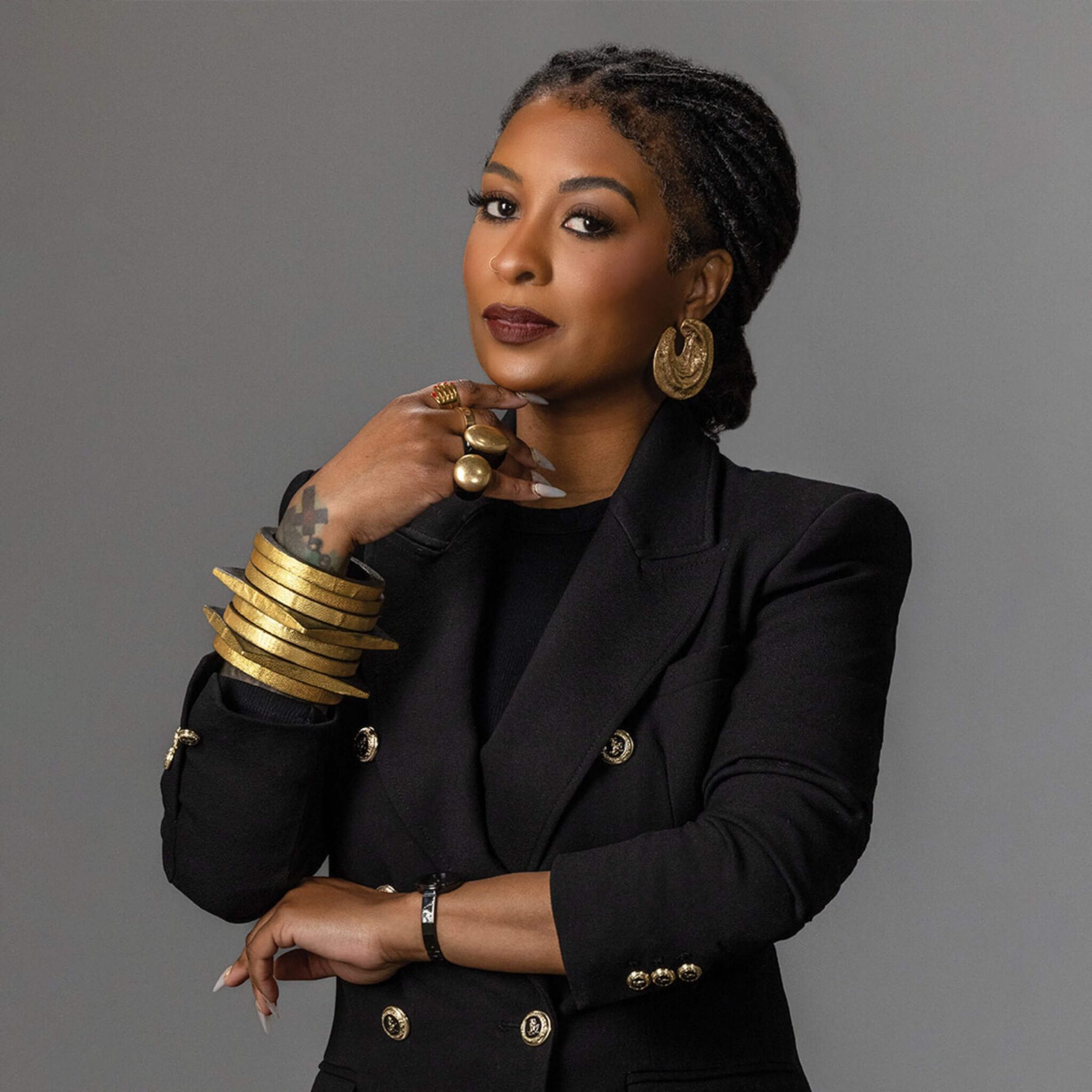 woman with gold bracelets and rings in a black jacket posing in front of a grey background