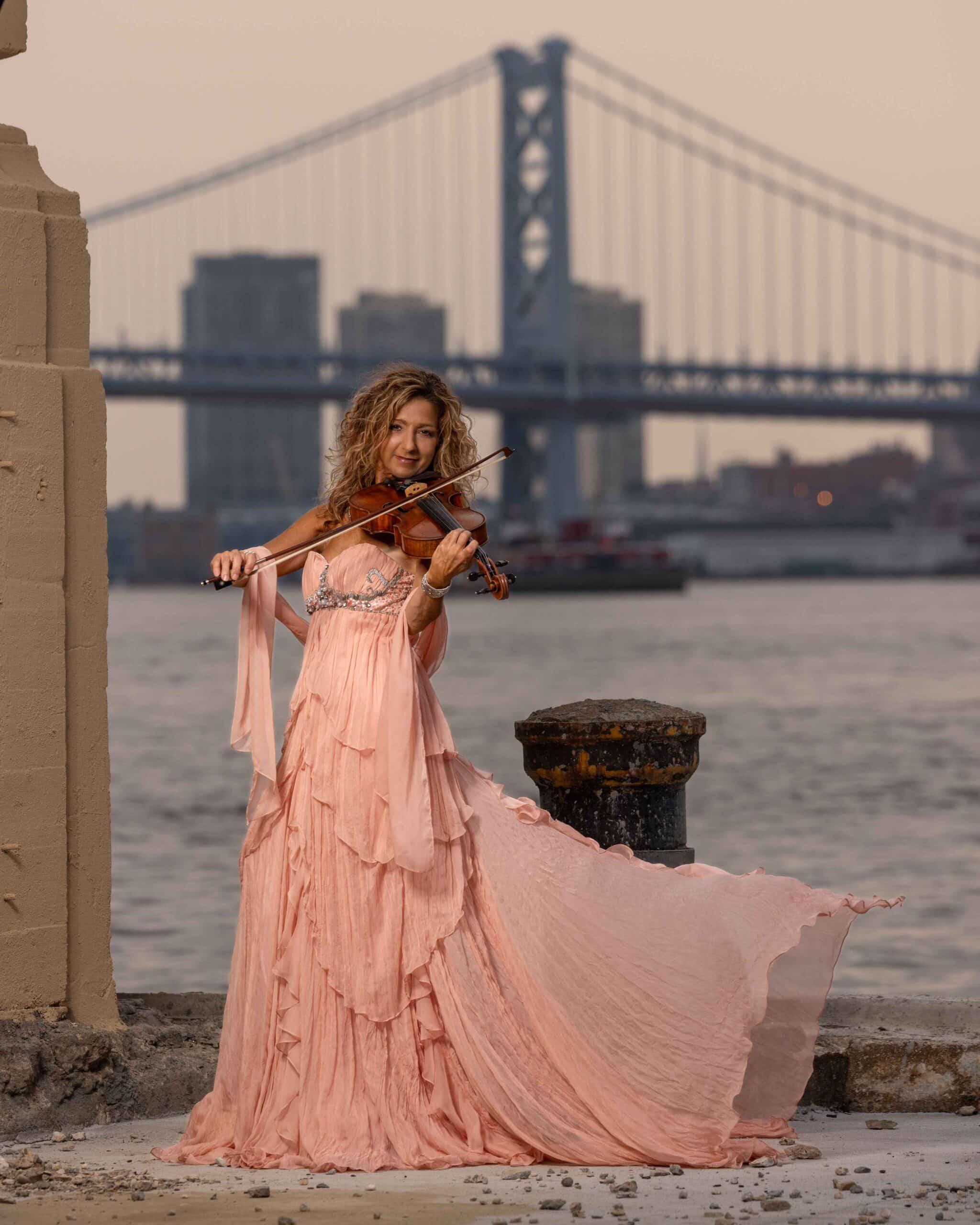 glamorous woman wearing a pink dress and playing the violin against a blue bridge skyline