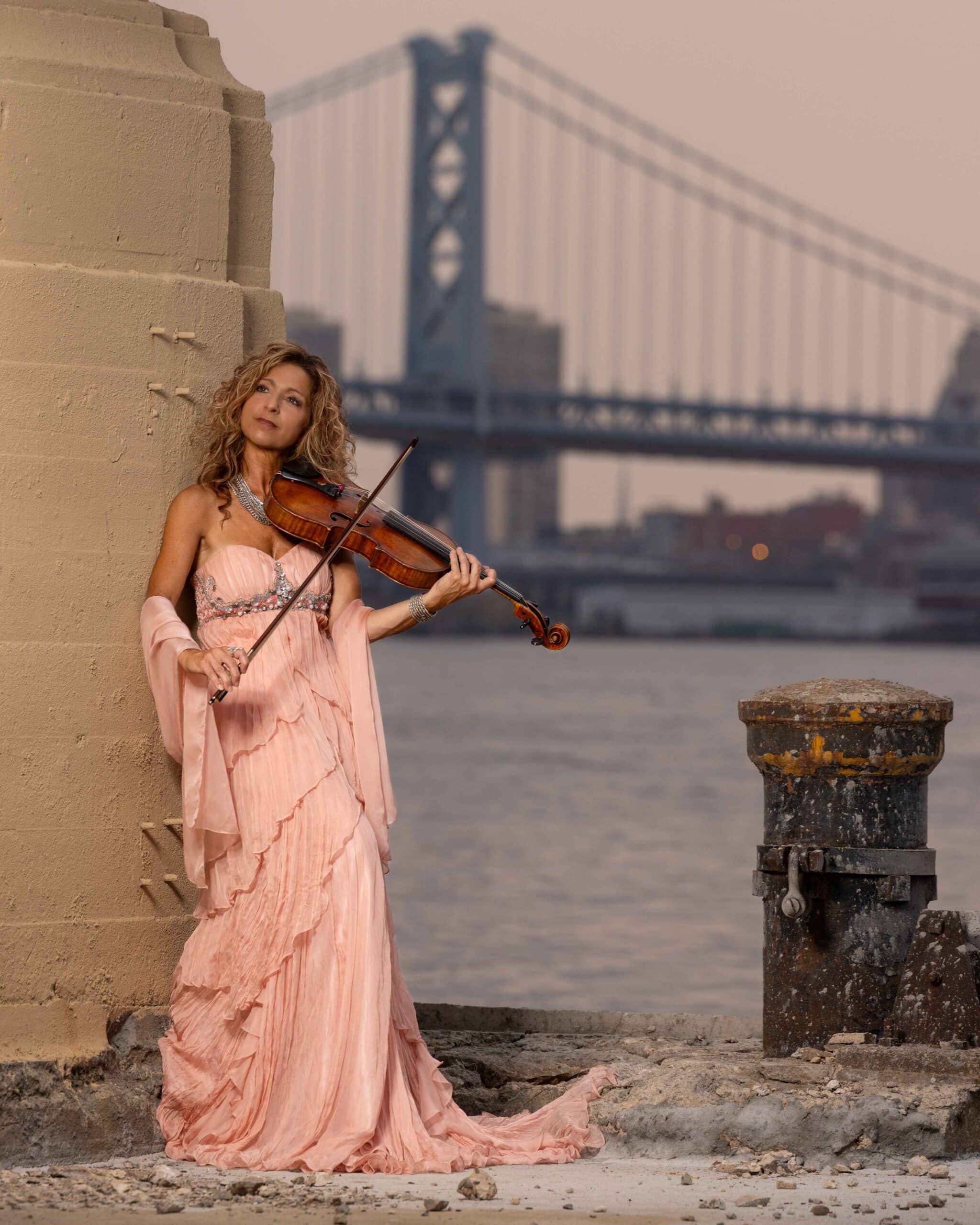 woman playing the violin while wearing a pink dress against a bridge skyline