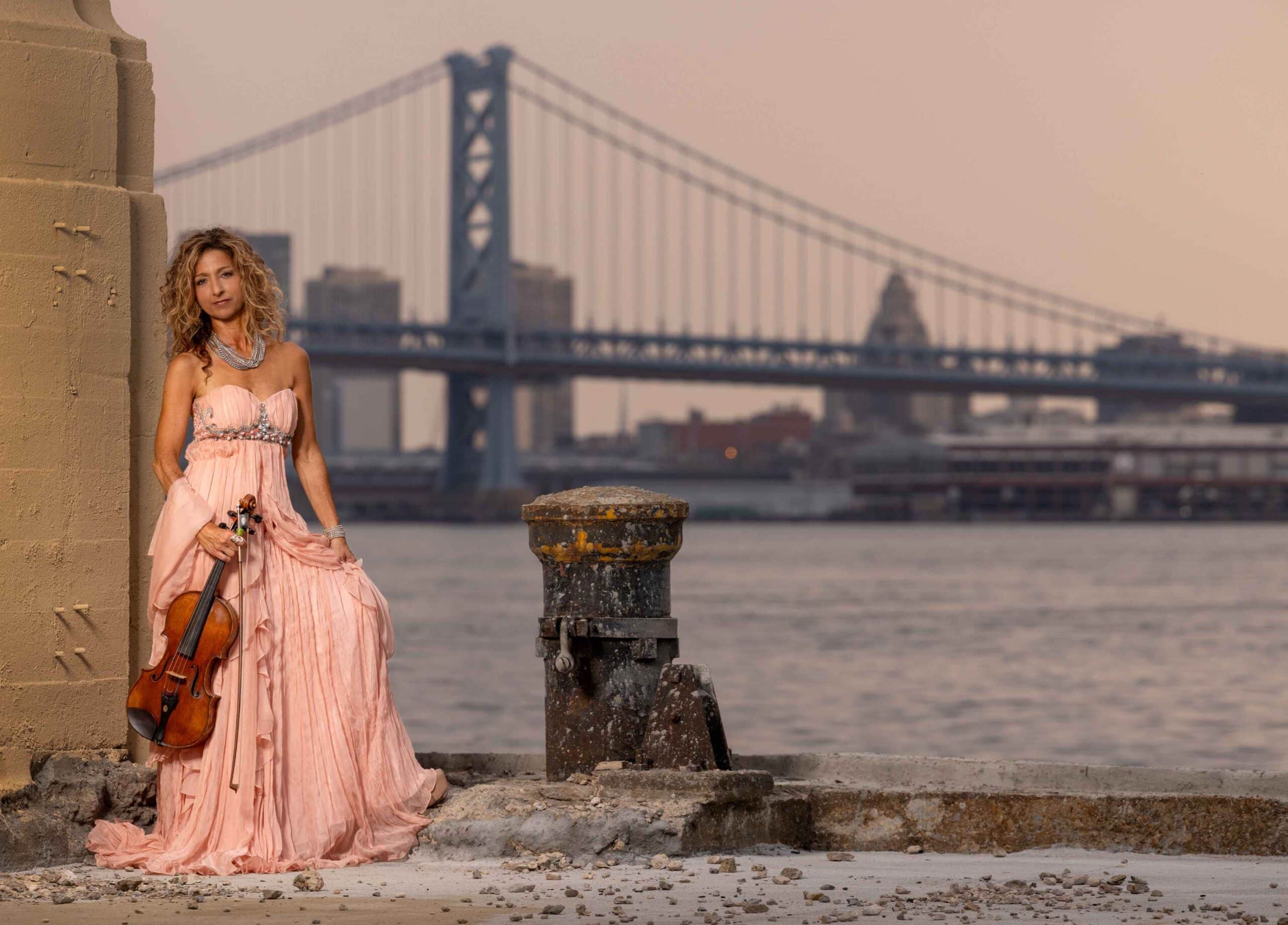 woman posing in a long pink dress while holding a wooden violin against a city skyline and bridge