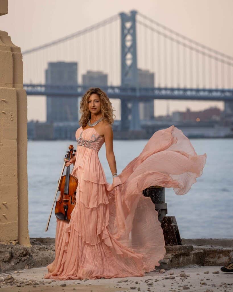 woman holding a violin and wearing a pink dress with bridge in background