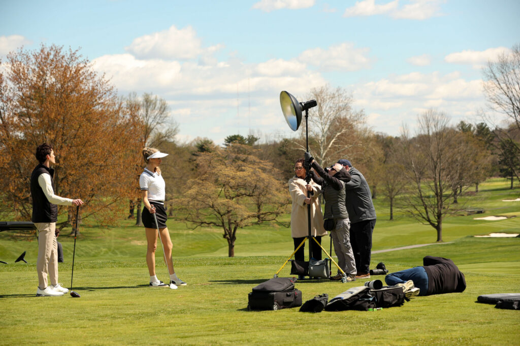 Production crew setting up lighting and camera equipment for a luxury golf fashion editorial with scenic golf course views.