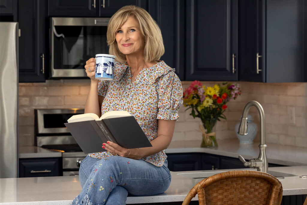 blonde hair woman sitting on kitchen counter reading a book and drinking from blue and white mug
