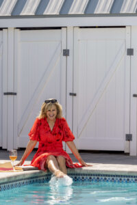 woman in red dress kicking the water in a swimming pool with white wine beside her in a glass