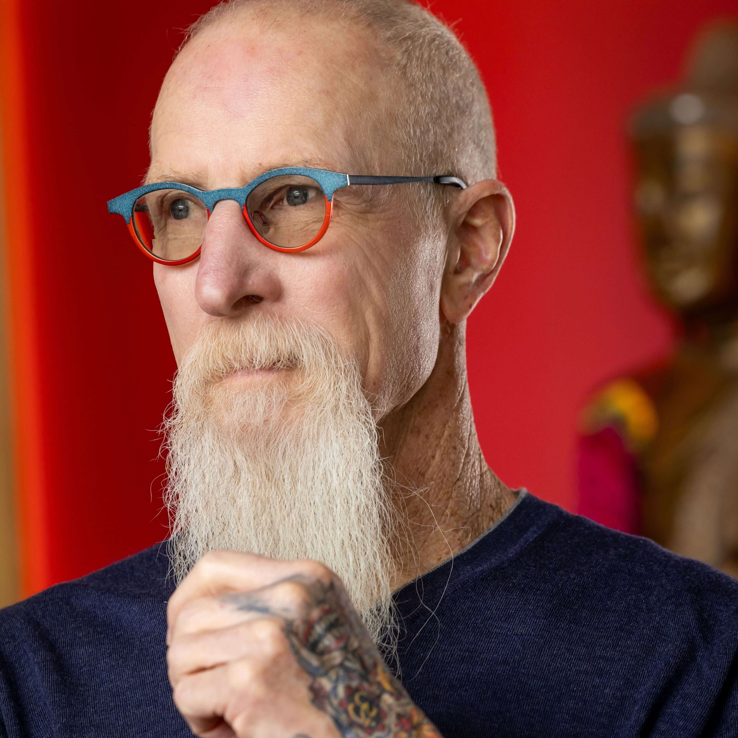man with white beard wearing blue and red glasses looking off camera against a red background