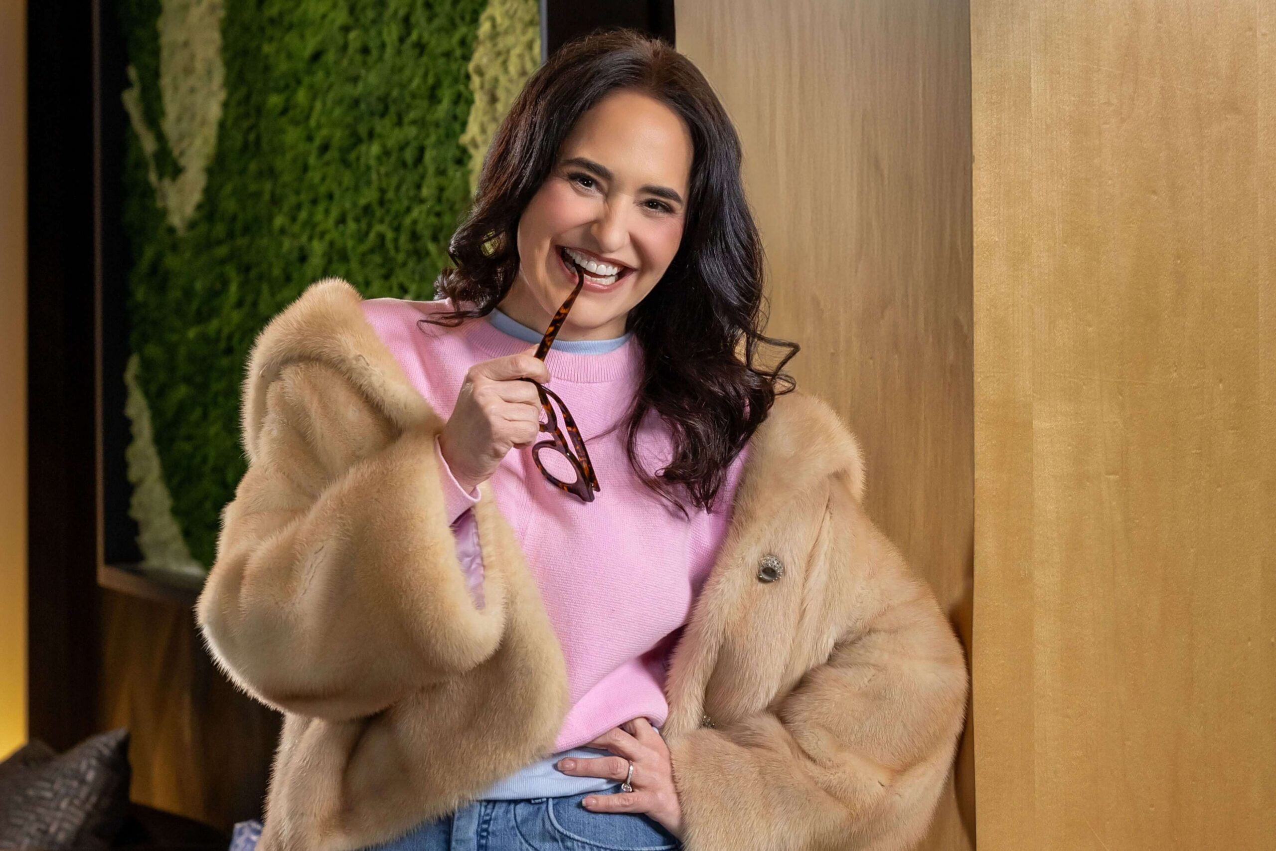 woman in a brown fur jacket and pink sweater biting glasses during editorial portrait photography session for a magazine. Photography by Phil Kramer