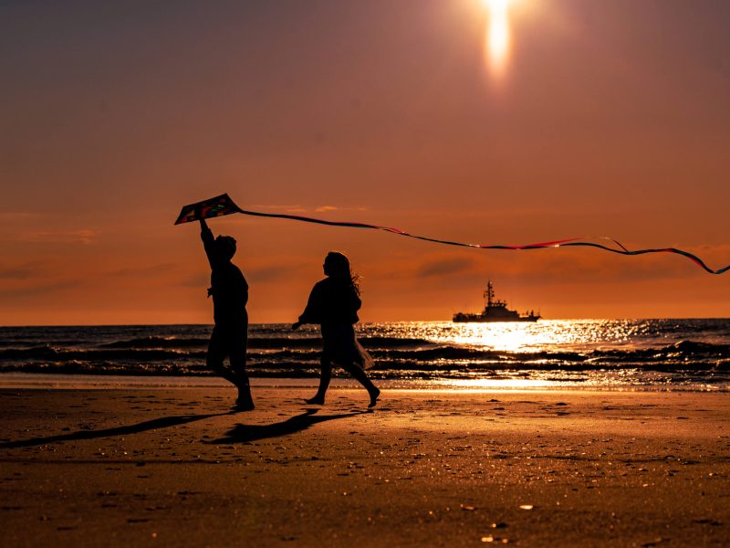 sunrise photo of children running on the beach with a kite, and boat in the background