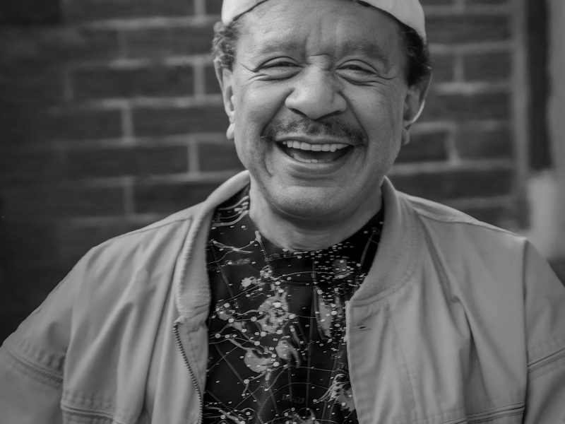 sherman hemsley smiling while wearing a white hat and khaki jacket against a brick wall